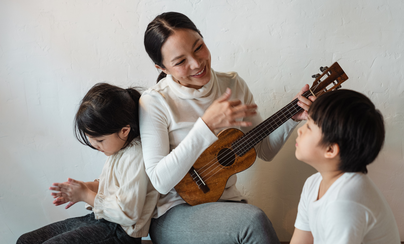 Smiling ethnic female playing ukulele while sitting with children
